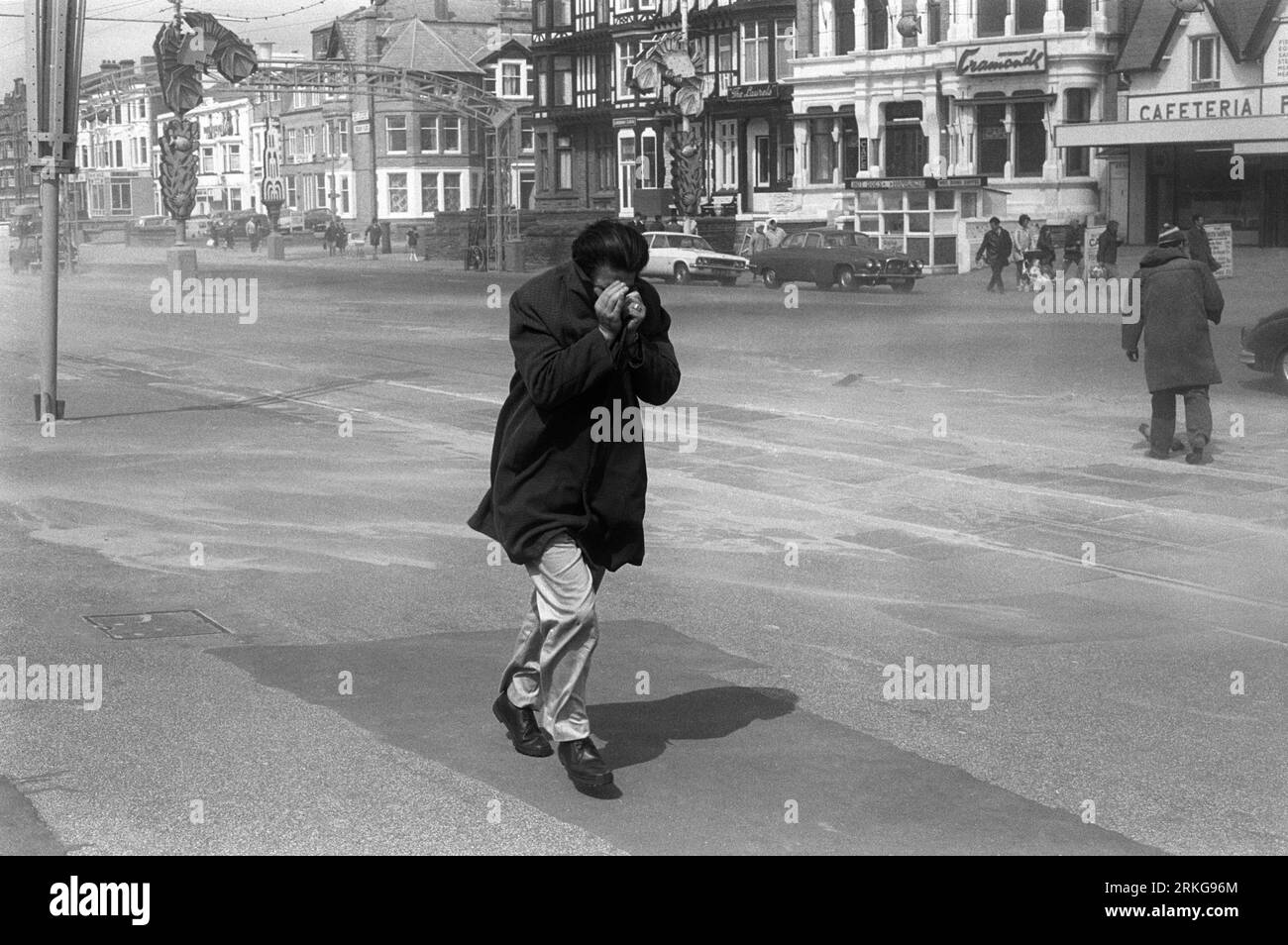 Golden Mile Blackpool 1970s UK. Ein starker Wind bläst trockenen Sand vom Strand und auf die Golden Mile Promenade. Ein Mann versteckt sein Gesicht in seinem Mantel gegen die brennenden, vom Wind geblasenen Sandpartikel. Blackpool, Lancashire, England um August 1970. HOMER SYKES Stockfoto