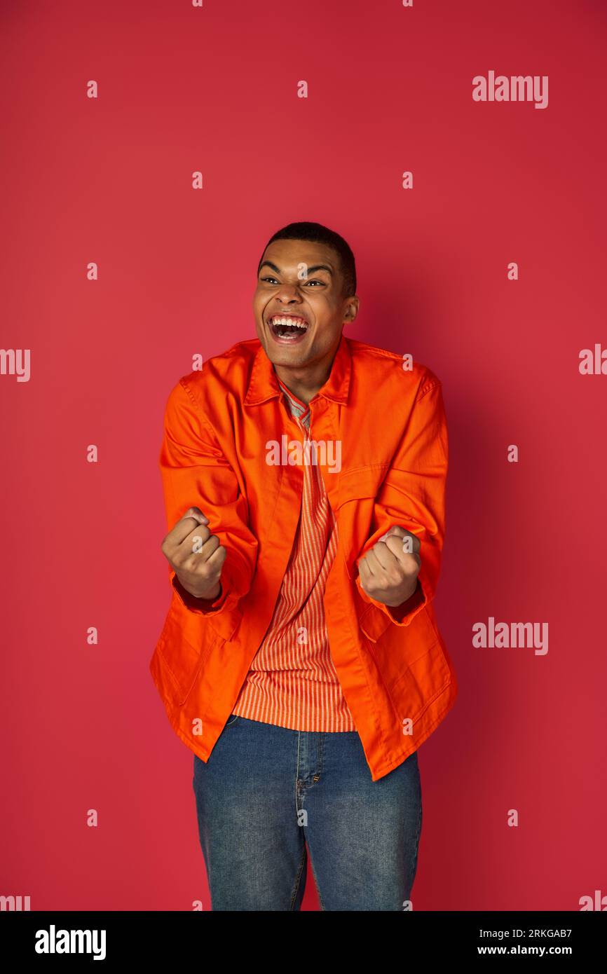 delighted, excited african american shouting and showing win gesture, orange shirt, red background Stockfoto
