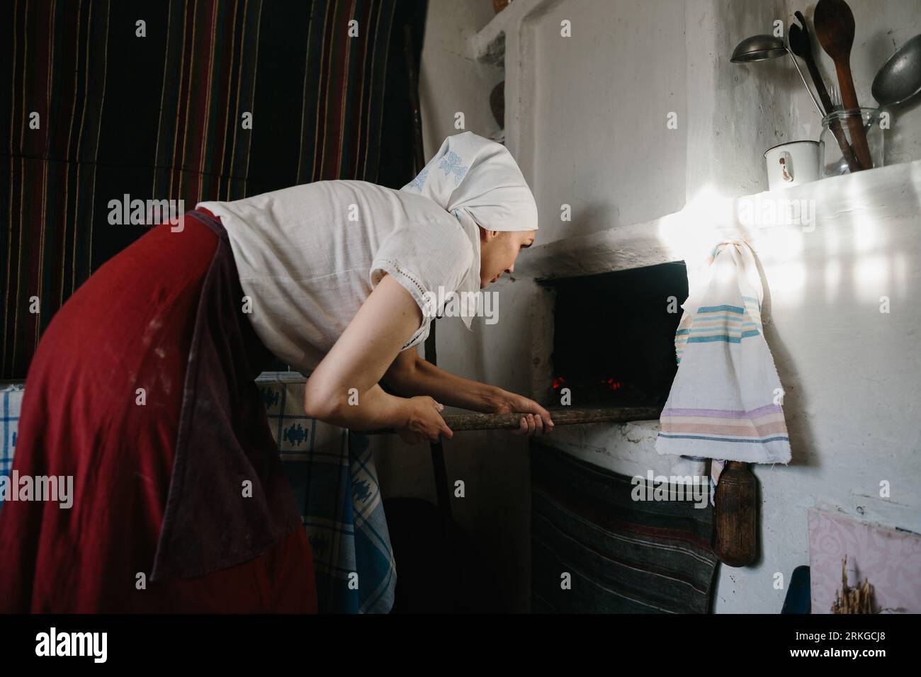 Eine Frau in ukrainischer Nationalkleidung arbeitet in der Nähe eines Holzofens. Der traditionelle Prozess des Brotbackens zu Hause. Stockfoto