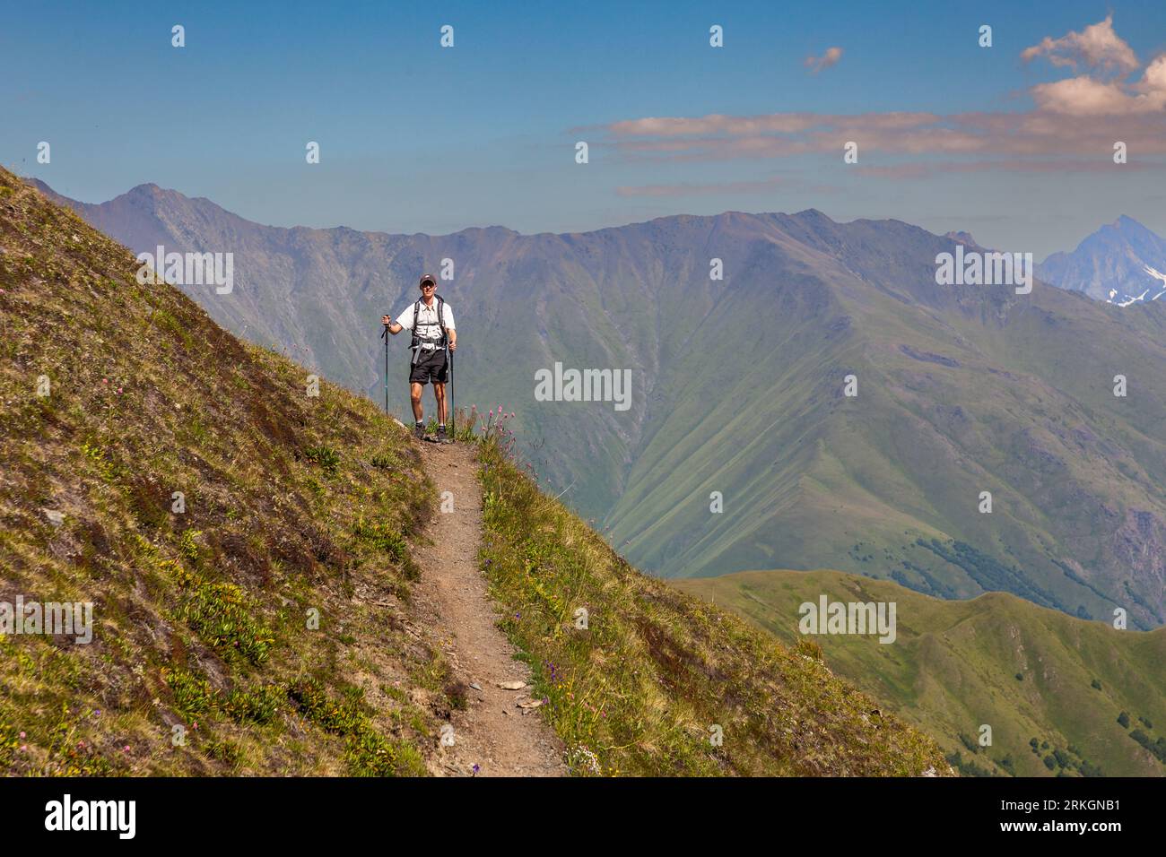 Julius Berg steht mit Trekkingstöcken auf einem schmalen Pfad während einer Wanderung im Hochkaukasus, auf dem Abstieg vom Atsunta-Pass auf der Khevsur-Seite nahe dem Dorf Andaki in der Gemeinde Duscheti, Region Mtskheta-Mtianeti, Georgien. Er trägt ein weißes Hemd, dunkle Shorts, eine Kappe, eine Sonnenbrille und einen Rucksack. Hinter ihm fällt der Hang in ein Tal ab. Ein grünes Gebirgszug erhebt sich darüber. Rechts ragt ein Gipfel mit Schneefeldern empor. Die Trekkingtour mit Packpferden umfasste fast 120 Kilometer und mehr als 5.000 Meter Auf- und Abstieg. Khevsureti (georgisch ხევსურეთი) ist eine historische Region am th Stockfoto
