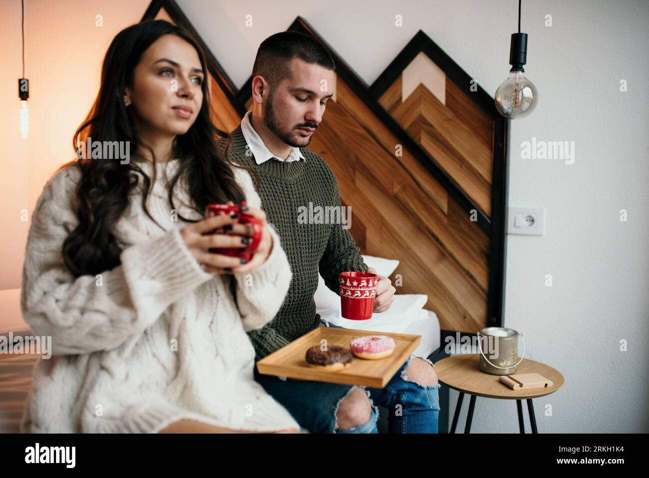 Ein locker gekleidetes Paar, das nahe beieinander auf einem Bett sitzt und beide eine Tasse Kaffee in der Hand hält Stockfoto