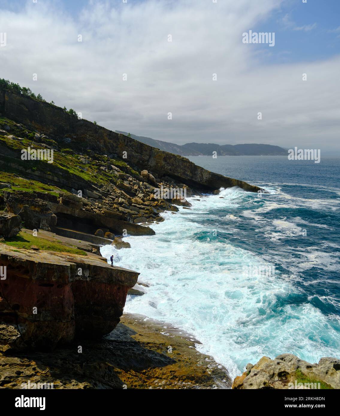 Vertikale Landschaft einer Küste mit einer Person, die das Meer von einem riesigen Felsen aus beobachtet, die Wellen treffen die Küste Stockfoto