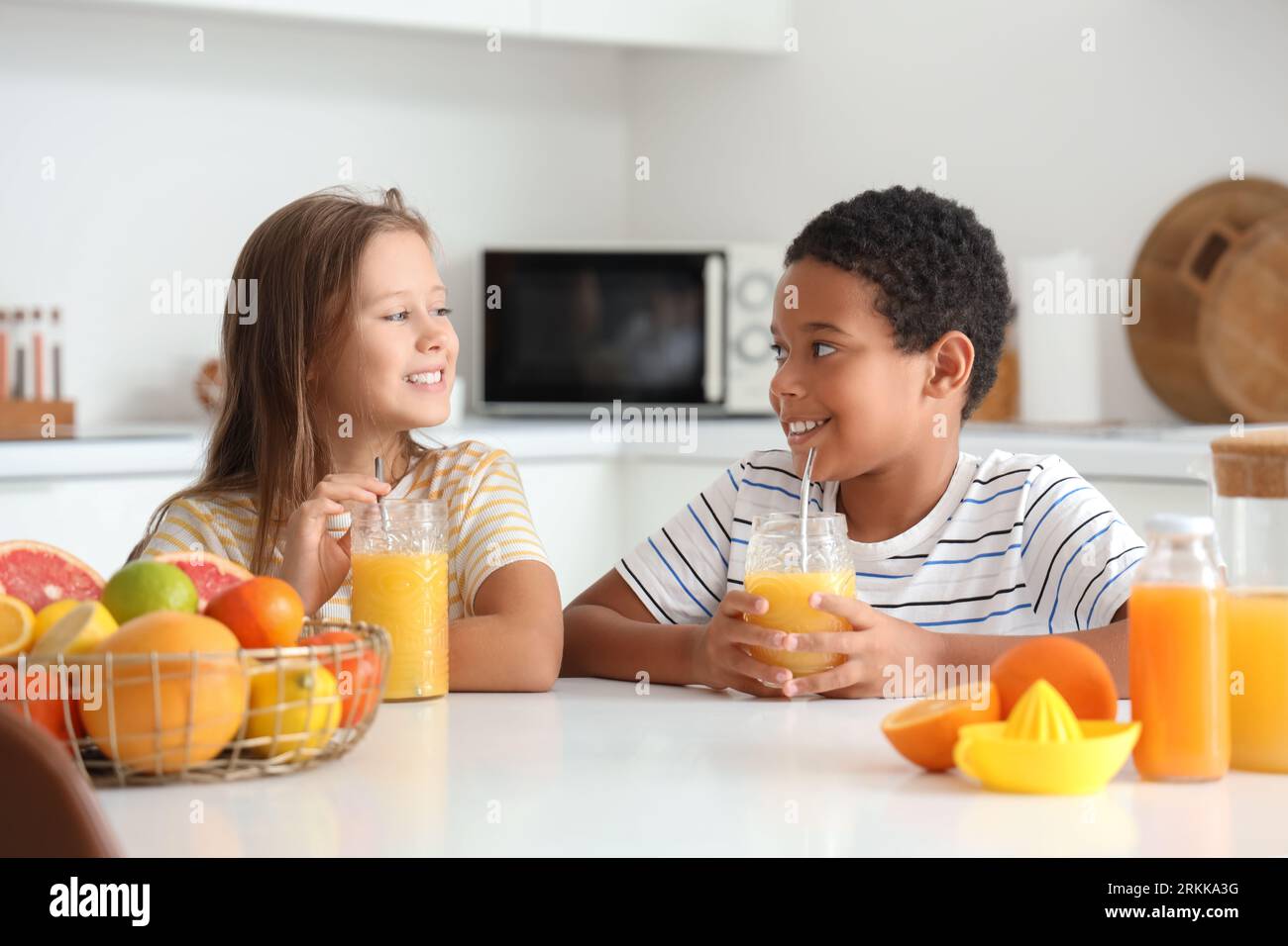 Little children with glasses of fresh citrus juice sitting at table in kitchen Stockfoto