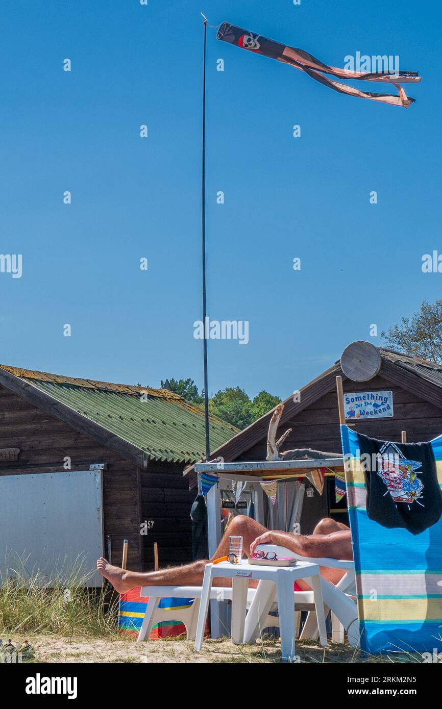 Zusammenfassung. Mann sonnt sich die Beine und entspannt sich vor einem typischen British Beach Hütten in Dorset. UK. Sommer. Sonnig. Keine Fläche wird angezeigt. Klassisch Britisch. Stockfoto