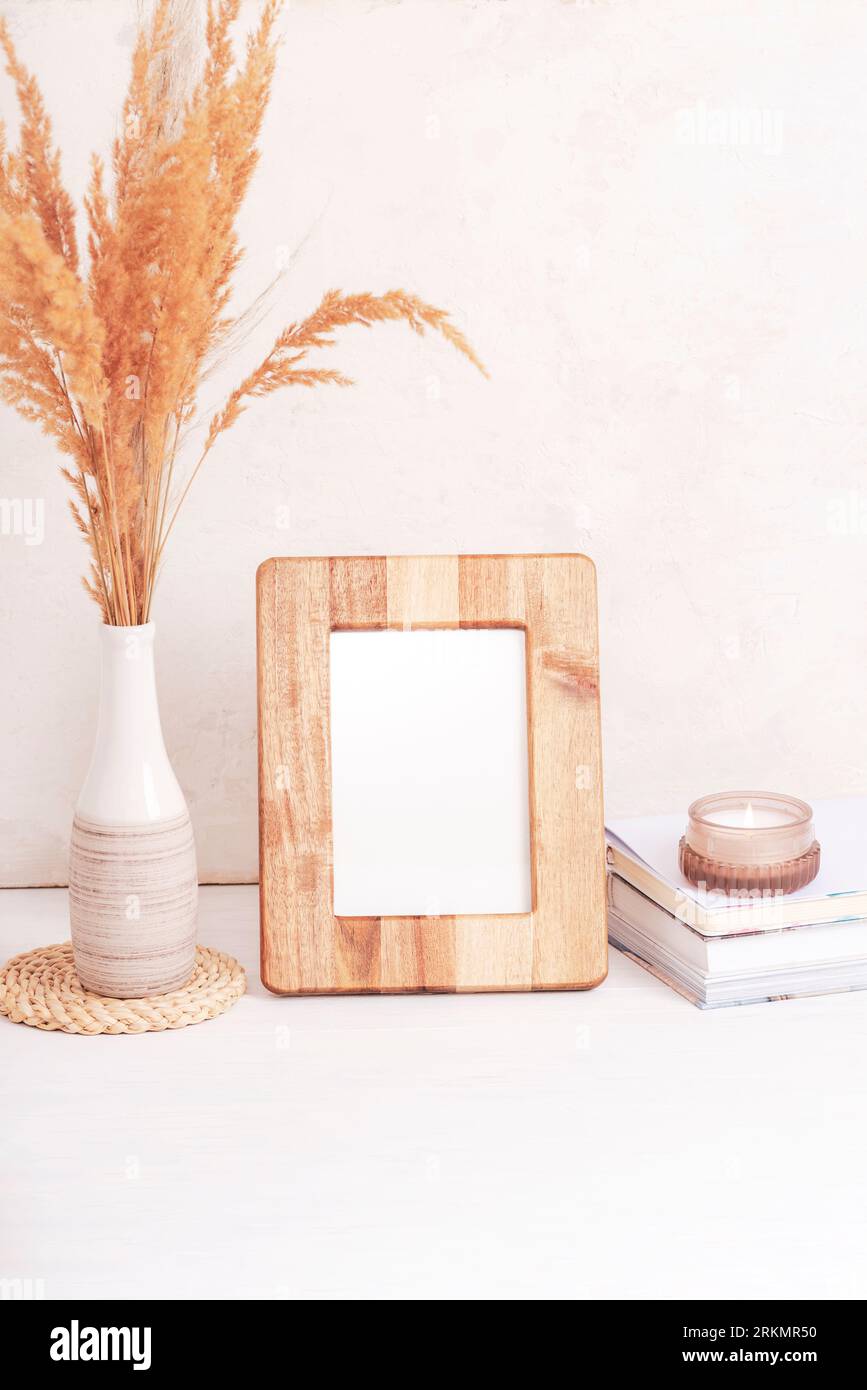 Mockup-Bilderrahmen aus Holz, Bücher, Duftkerze und Pampasgras in Vase auf weißem Tisch. Leerzeichen kopieren. Stockfoto