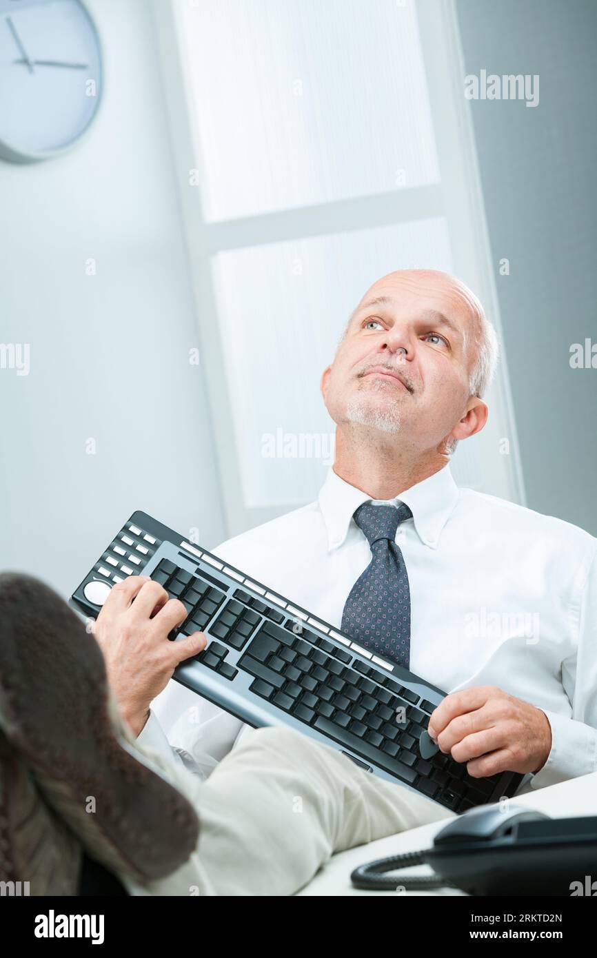 In einem Büro gibt ein leitender Geschäftsmann freudig vor, eine Computertastatur als Gitarre zu spielen. Mit den Füßen auf dem Tisch träumt er Tagträume und verkörpert den spiri Stockfoto