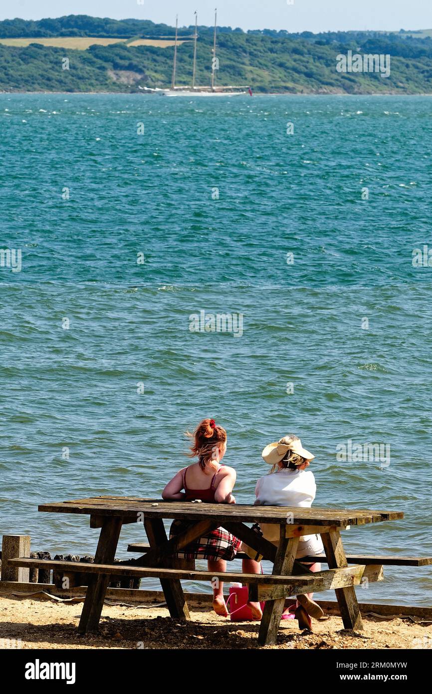Zwei junge Frauen sitzen auf einer Tischbank am Strand von Lepe und blicken an einem heißen Sommertag über den Solent zur Isle of Wight in Hampshire England Stockfoto