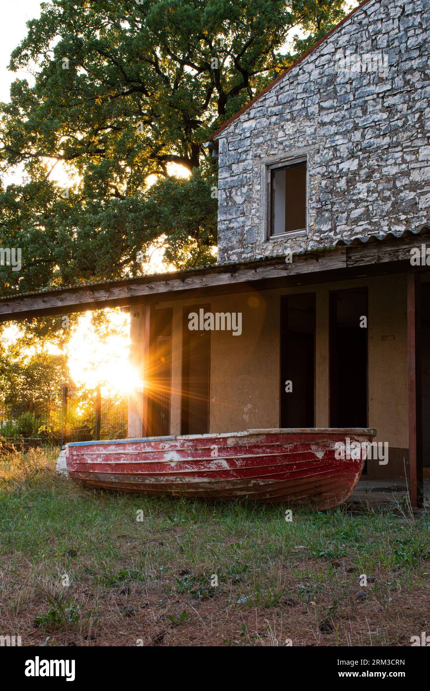Sonne scheint durch die Bäume auf dem kleinen roten Holzboot vor einem Haus aus Stein in Umag, Kroatien. Stockfoto