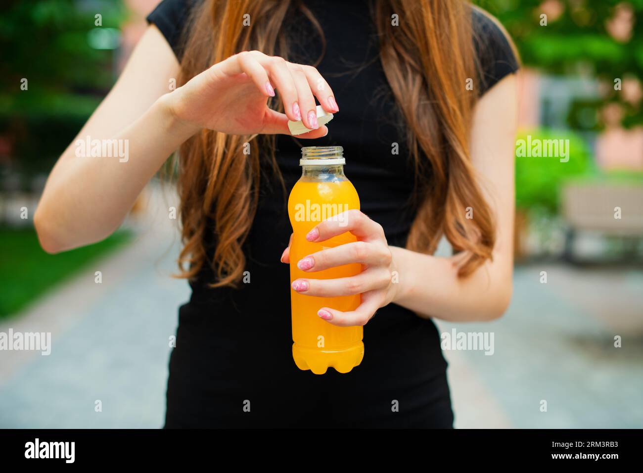 Eine nicht erkennbare junge Frau öffnet an einem Sommertag draußen eine orange Limonade-Plastikflasche, die in einer Stadtstraße in bunten Gebäuden steht Stockfoto