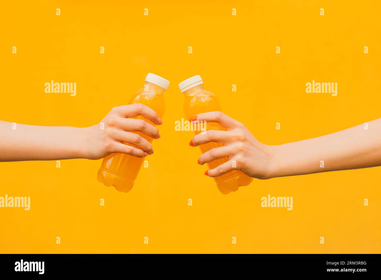 Zwei nicht erkennbare junge Frauen halten in seiner Hand die orange Limonade Plastikflasche auf gelbem Straßenmauerhintergrund, Nahaufnahme Seitenansicht Stockfoto