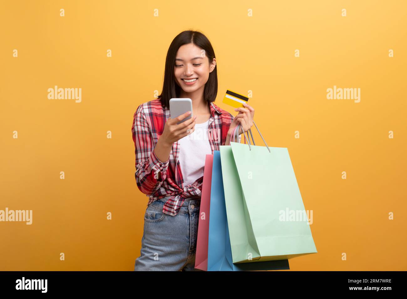 Coole lächelnde asiatische Dame in lässiger Haltung mit bunten Einkaufstaschen, Smartphone und Bankkarte, gelber Studiohintergrund Stockfoto