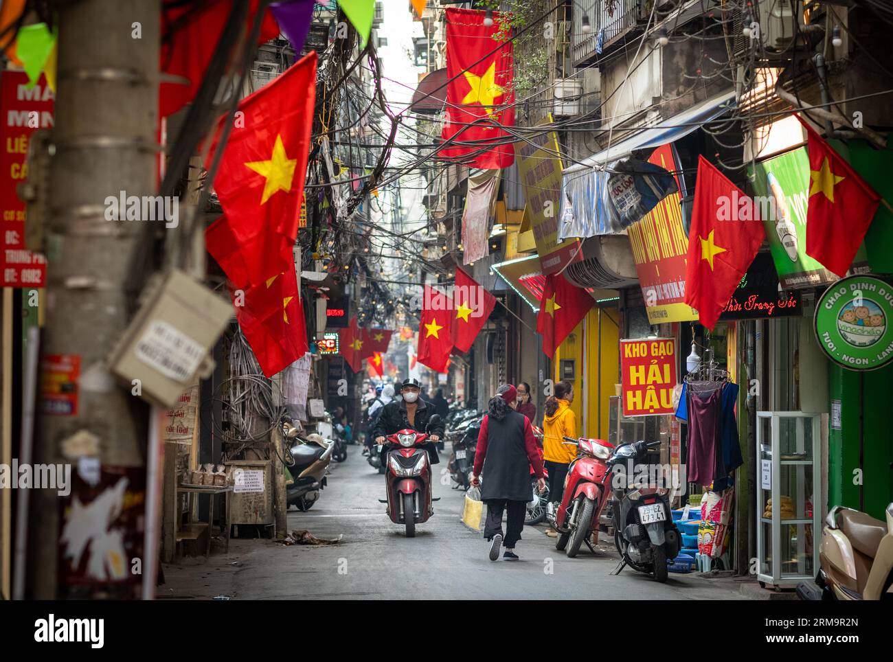 Die Menschen gehen unter vietnamesischen Flaggen in einer engen Gasse namens Kham Thien Market in Hanoi, Vietnam, über das tägliche Leben. Stockfoto
