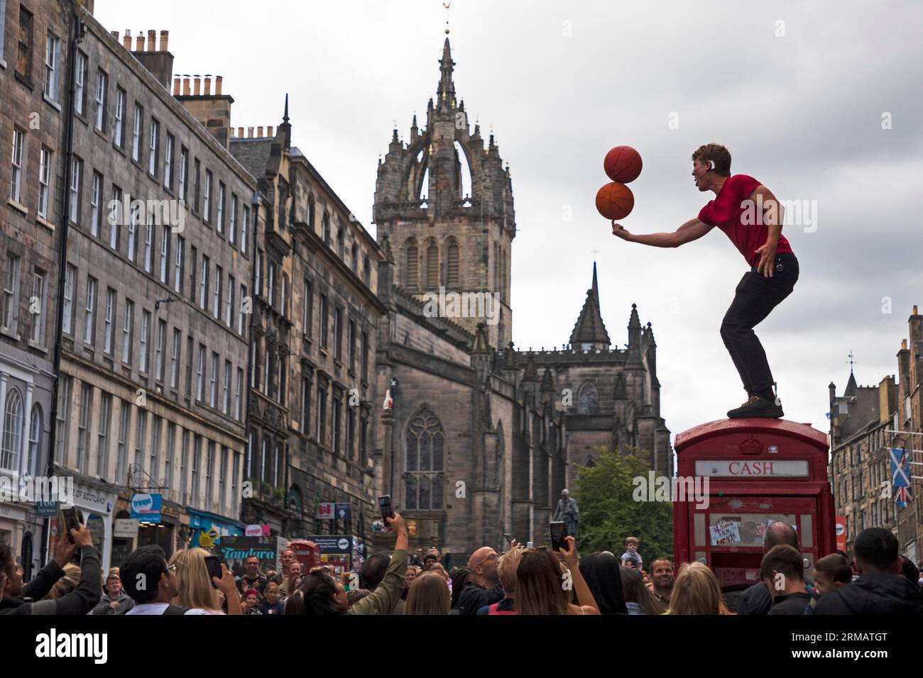 Royal Mile, Edinburgh, Schottland, Großbritannien. 27. August 2023. Obwohl das offizielle Edinburgh Festival Fringe am Montag, den 28. August endet, gab es auf der High Street keine Sicherheits- oder Fringe-Mitarbeiter, um die Massen und die Zeit der Straßenkünstler zu überwachen. Es gab jedoch immer noch große Zuschauergruppen, die sich zur Unterhaltung versammelten. Hunter aus Hawaii demonstriert seine Fähigkeit, Basketbälle auf seinem Finger auszubalancieren, während er auf einem ehemaligen Telefonkiosk sitzt. Quelle: Arch White/Alamy Live News. Stockfoto