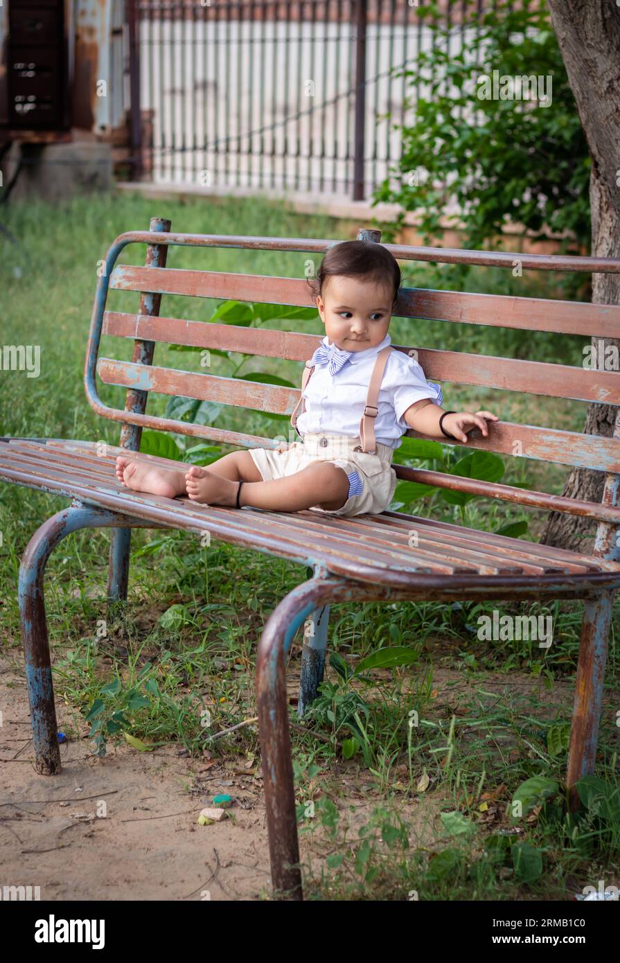 Isoliertes, süßes Kleinkind mit unschuldigem Gesichtsausdruck, das draußen auf einer rostigen Eisenbank sitzt Stockfoto