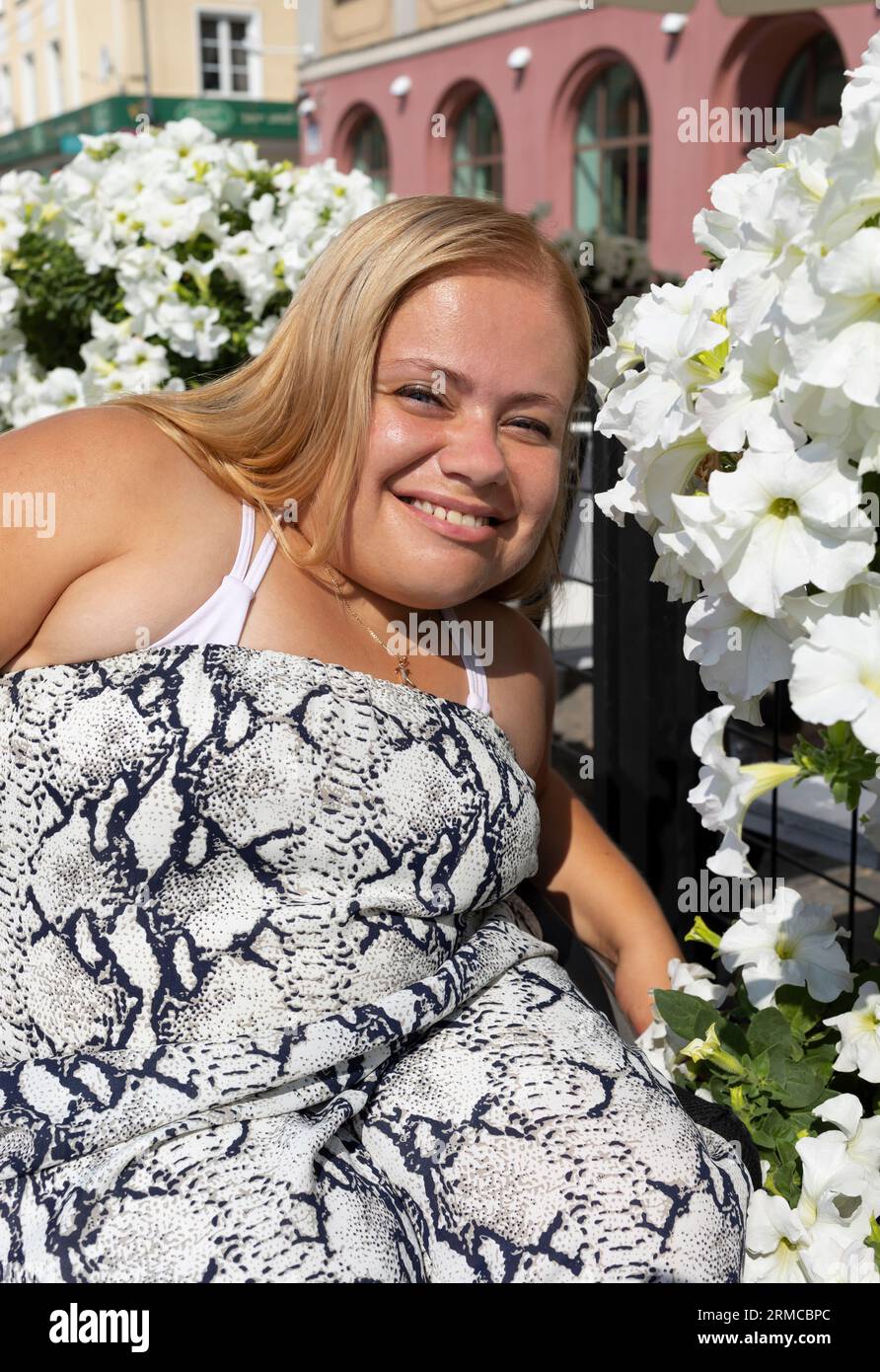 Junge Lächelnde Frau Mit Kurzem Körperbau Auf Rollstuhl, Die Am Sommertag In Der Nähe Von Followern Posiert. Porträt einer erwachsenen Frau mit körperlicher Behinderung Stockfoto