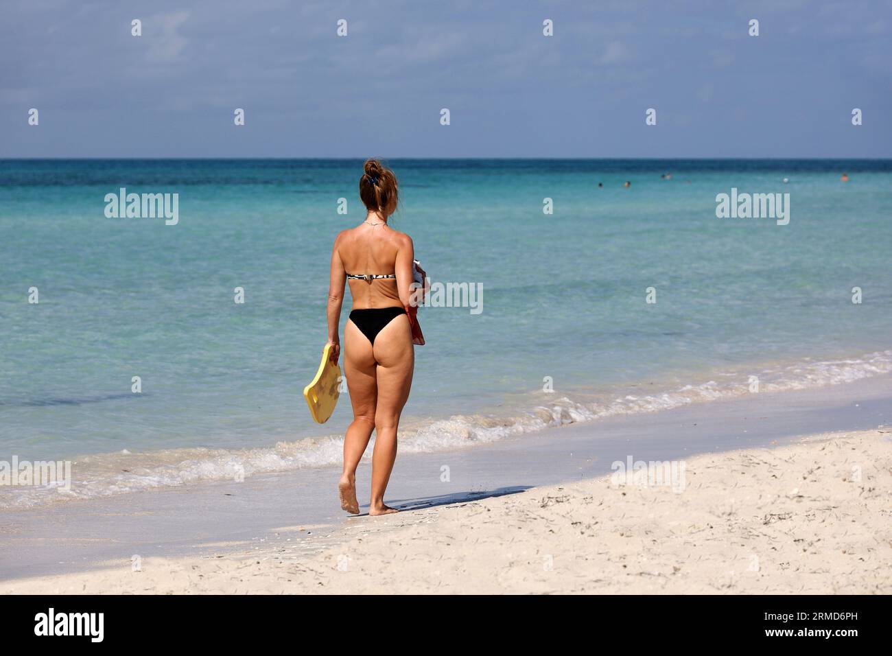 Junge Frau in schwarzem Bikini, die durch einen Sand auf blauem Meerwasserhintergrund geht. Strandurlaub an der Küste Stockfoto
