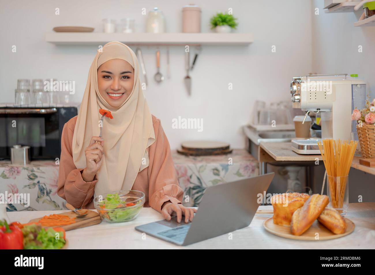 Eine schöne asiatische Muslimin isst ihren gesunden Salat, während sie auf ihrem Laptop an einem Esstisch in der Küche arbeitet. Arbeiten von zu Hause aus, freiberuflich, r Stockfoto