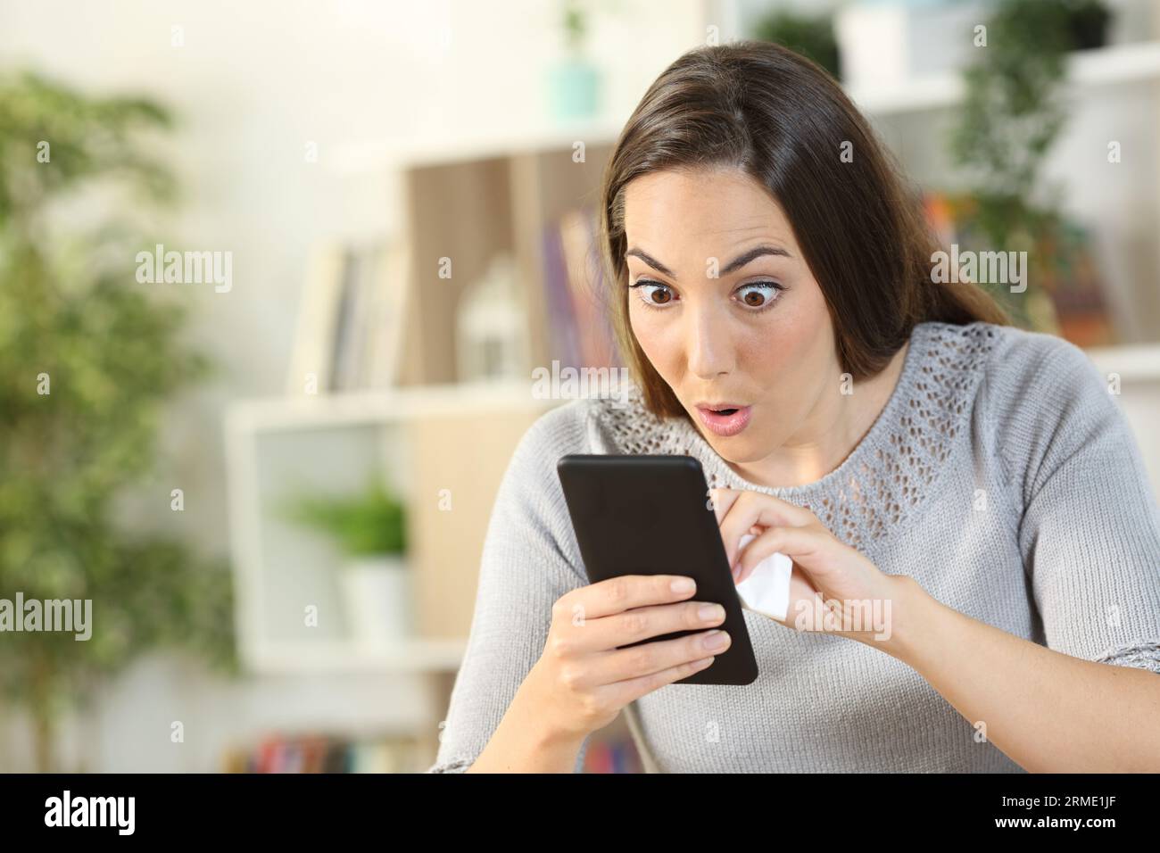 Überraschte Frau, die das Smartphone mit Augen und Mund aufputzte und zu Hause im Wohnzimmer saß Stockfoto