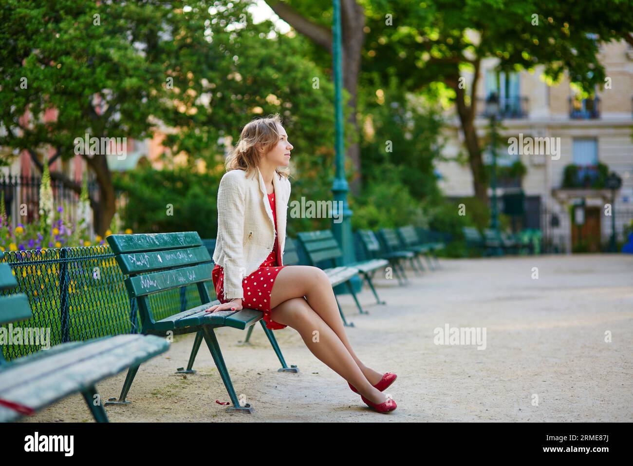 Schöne junge Frau in rotem Polka-Punkt-Kleid sitzt auf Bank in Marais, Paris, Frankreich Stockfoto