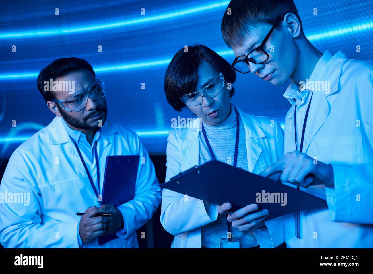 Junge Praktikantin, die auf das Clipboard in der Nähe multiethnischer Kollegen im Wissenschaftszentrum zeigt, zukunftsorientiert Stockfoto