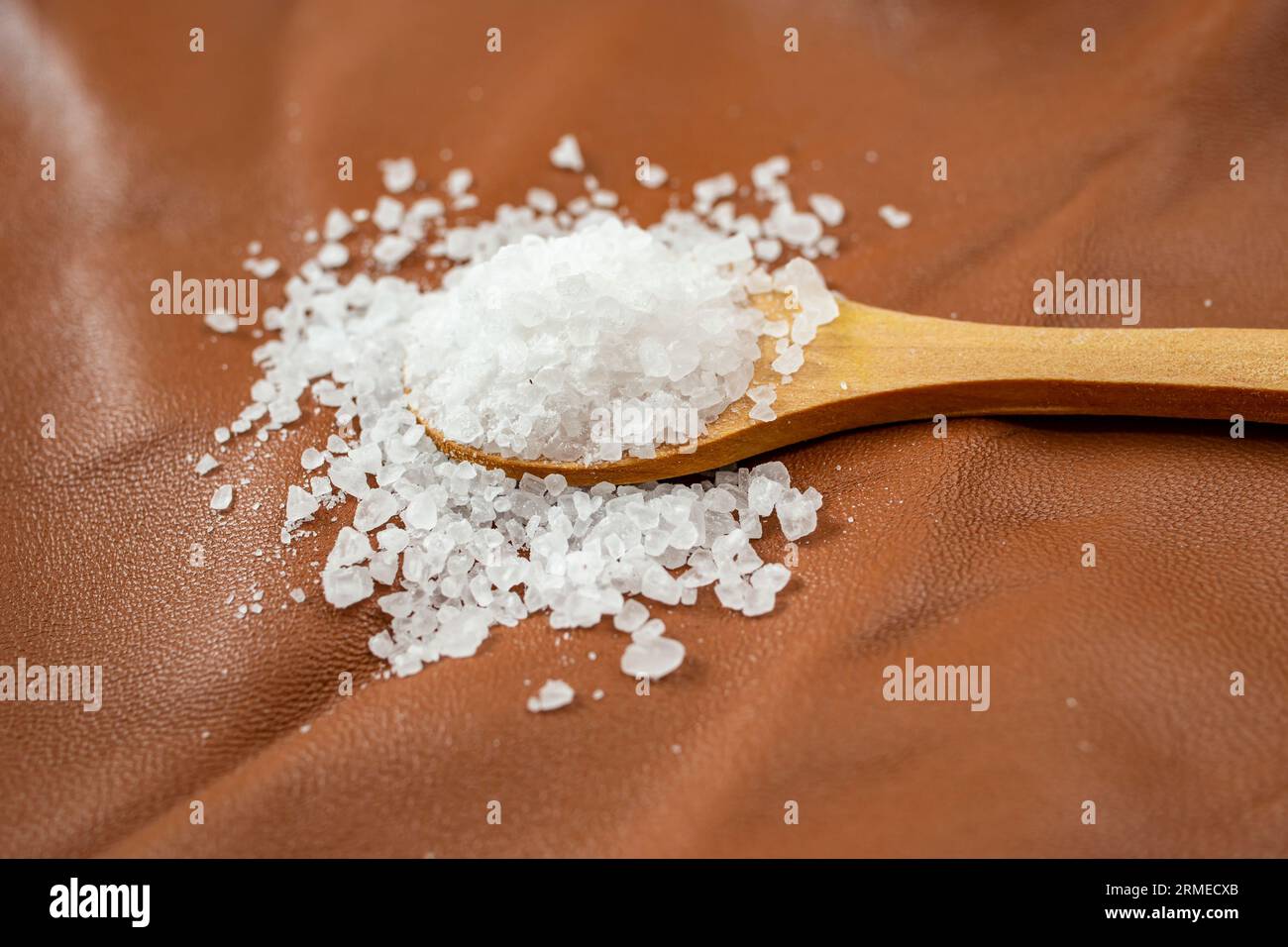 Weißes Steinsalz, gefüllt mit einem Teelöffel aus Holz, auf braunem Leder, Hauttextur, Seitenansicht, abstrakter Hintergrund mit weichem Fokus Stockfoto