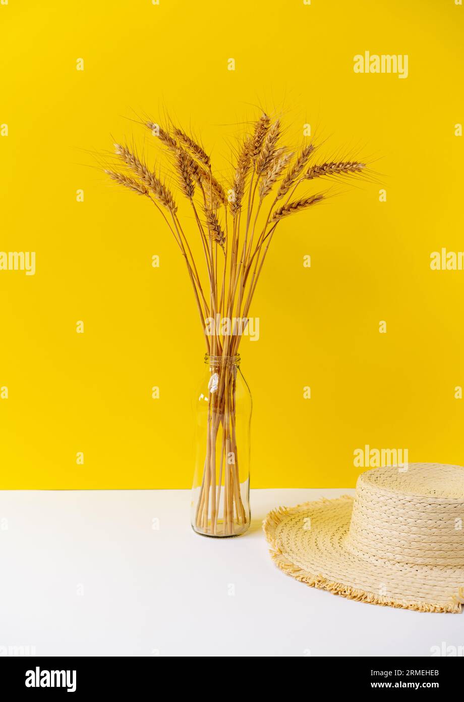 Stillebenstrauß aus gelbem Weizen in Vase aus Glasflasche und Strohsonnenhut auf weißem Tisch vor gelbem Hintergrund. Stockfoto