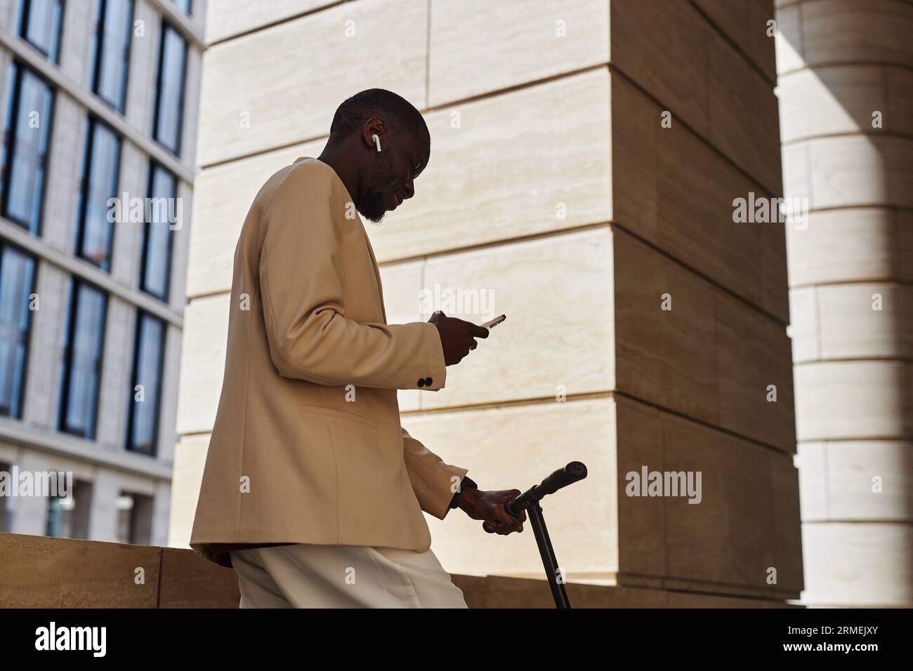 Seitenansicht eines jungen afroamerikanischen Geschäftsmannes mit Kopfhörern und Anzug, der sich im Mobiltelefon die Playlist anschaut, während er am Gebäude steht Stockfoto
