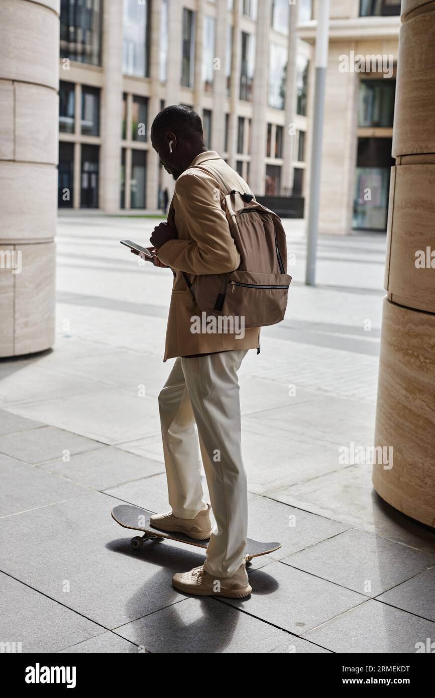 Rückansicht eines jungen eleganten Mannes mit Rucksack und Smartphone, der den Fuß auf dem Skateboard hält, während er vor der Kamera steht und SMS schreibt Stockfoto