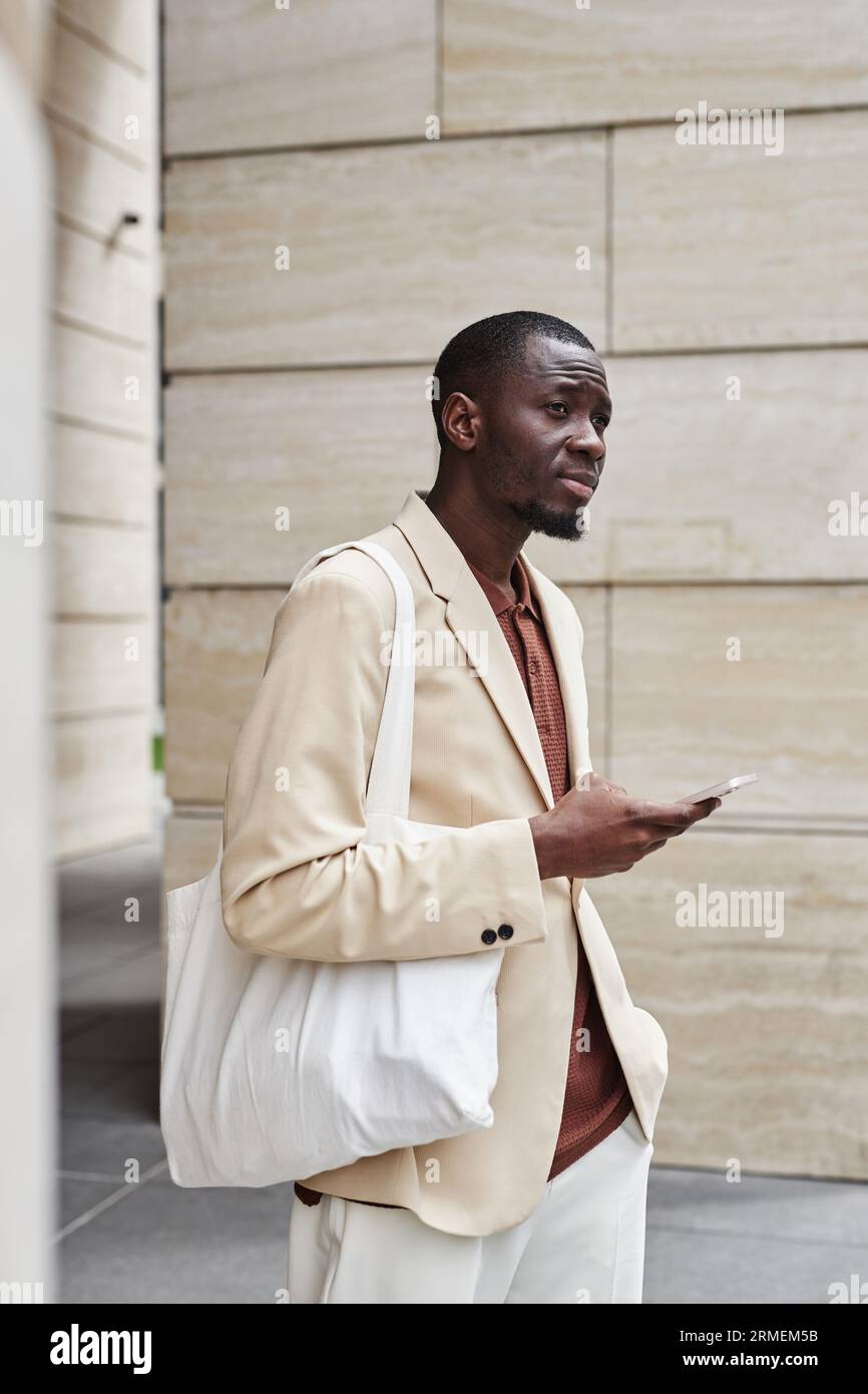 Junger, ruhiger Geschäftsmann mit weißer Textilhandtasche auf der Schulter, der auf Kollegen wartet und Smartphone benutzt, während er vor der Kamera steht Stockfoto