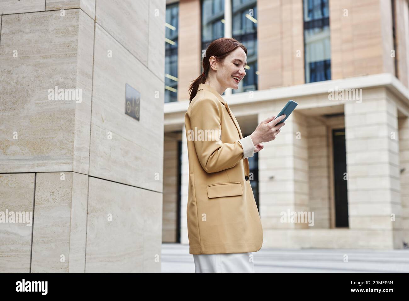 Seitenansicht einer glücklichen jungen Geschäftsfrau in ruhiger Luxuskleidung, die auf den Smartphone-Bildschirm schaut, während sie an der Säule moderner Architektur steht Stockfoto