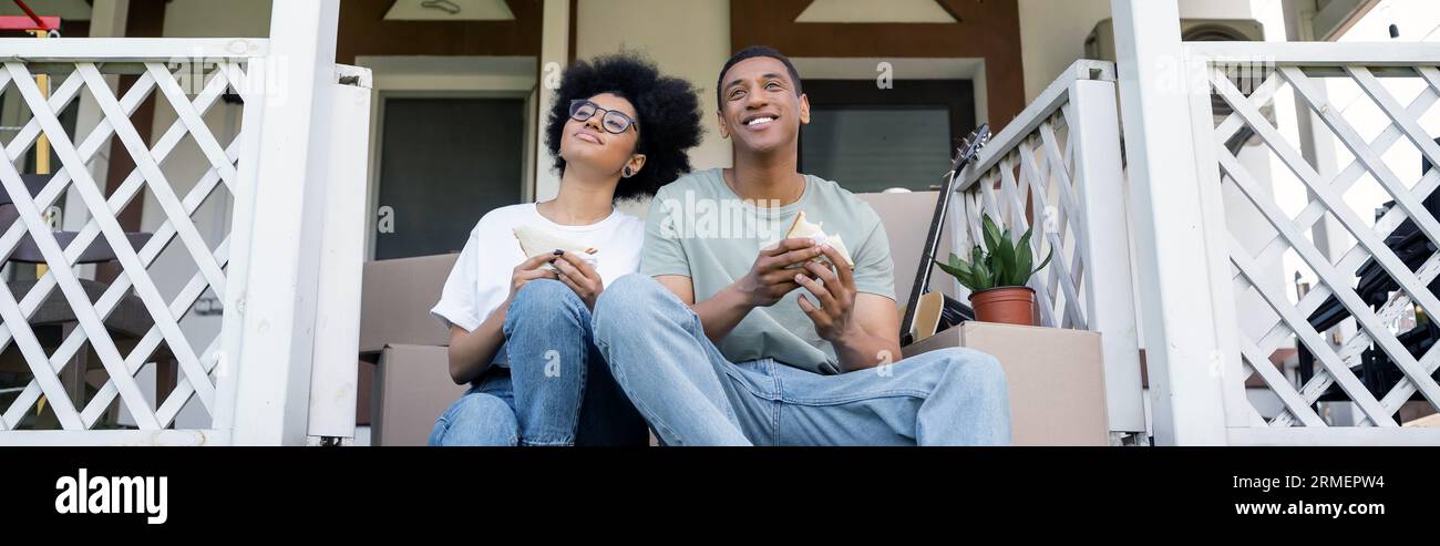 Zufrieden mit dem afroamerikanischen Paar, das leckere Sandwiches auf der Veranda des neuen Hauses, Immobilien, Banner hält Stockfoto