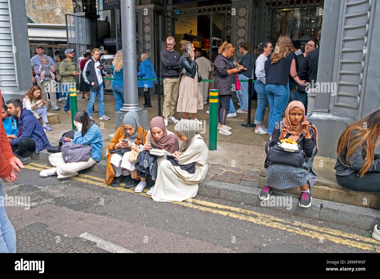 Muslimische Mädchen Jugendliche junge Frauen, die auf der Bordsteinkante sitzen und Straßenessen vor dem Borough Market Stoney St. essen South London SE1 England UK KATHY DEWITT Stockfoto