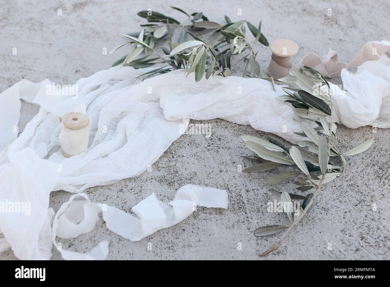 Mediterranes Hochzeitstillleben. Olivenbaumzweige, Früchte mit Seidenbändern und weißer Musselin-Tischkanne, Tuch oder Schleier. Grunge Beton Stockfoto