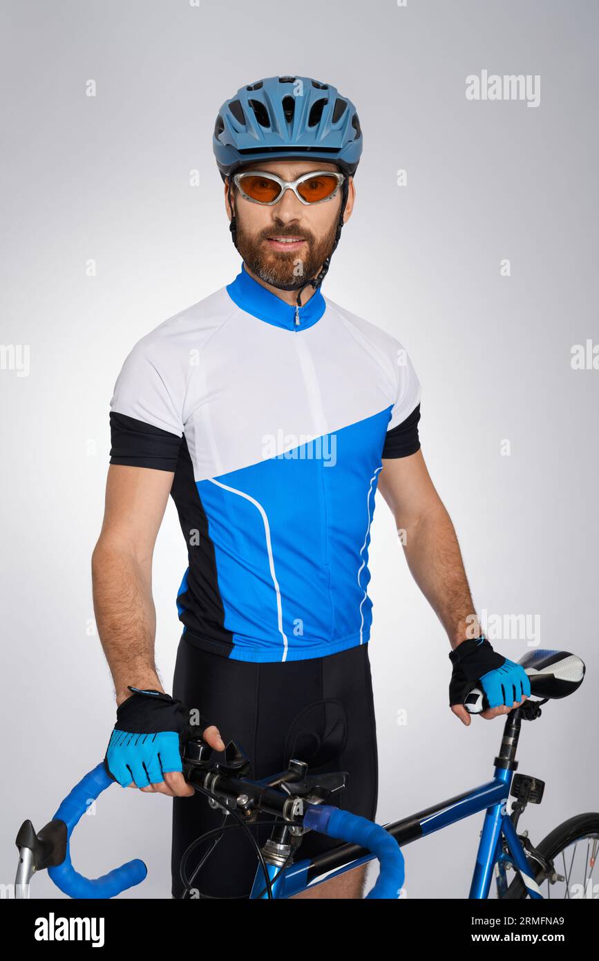 Professioneller Radfahrer im vollen Gang, mit Fahrrad im Studio stehend. Porträt eines lächelnden bärtigen Athleten in Schutzhelm und Brille, vor der Kamera posierend, isoliert auf Grau. Konzept des Radfahrens. Stockfoto