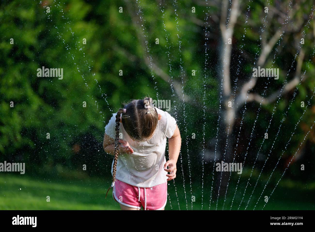 Ein junges Mädchen, das das warme Wetter im Garten unter dem Wassersprenger genießt Stockfoto