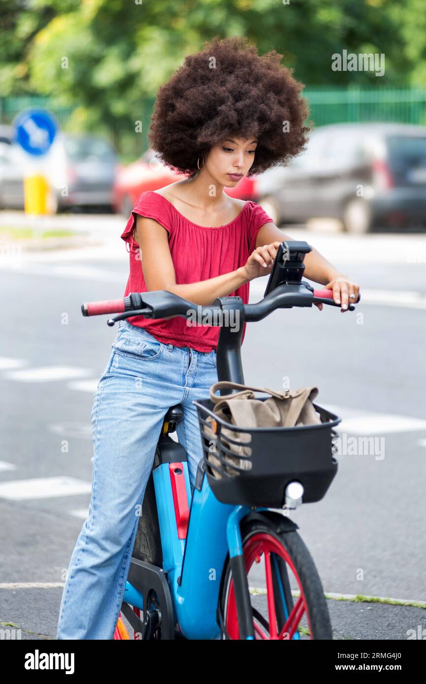 Stilvolle junge gemischte Rennfrau mit dunklem Afro-Haar in lässigem Outfit mit Smartphone, während sie auf der Asphaltstraße in der Stadt auf sonnigem da mit dem Elektrofahrrad fährt Stockfoto