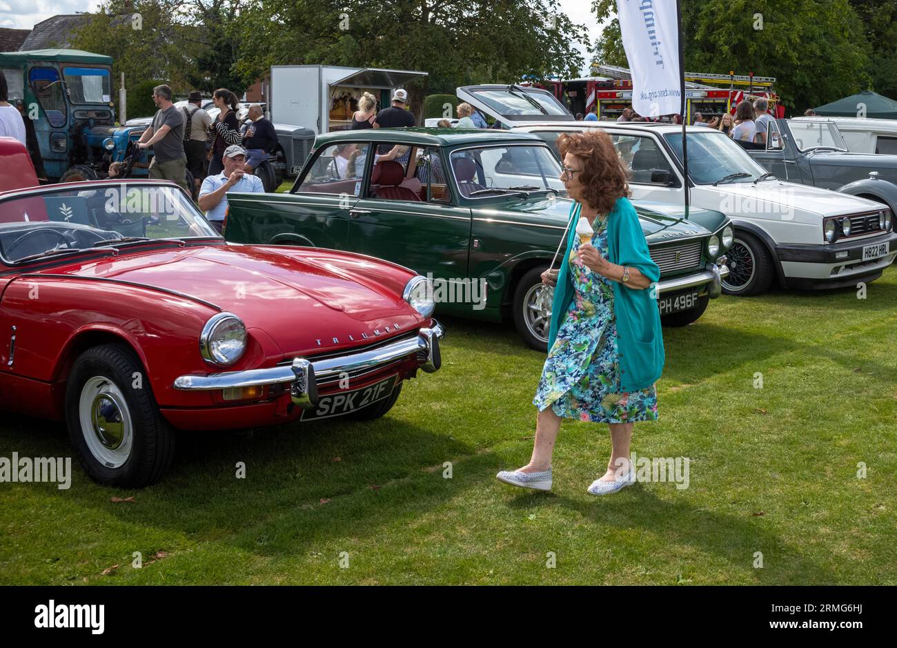 Eine Frau mittleren Alters hält ein Eis, während sie an klassischen und Oldtimern vorbeigeht, die im Wisborough Green Village Fete, West Sussex, Großbritannien, gezeigt werden. Stockfoto