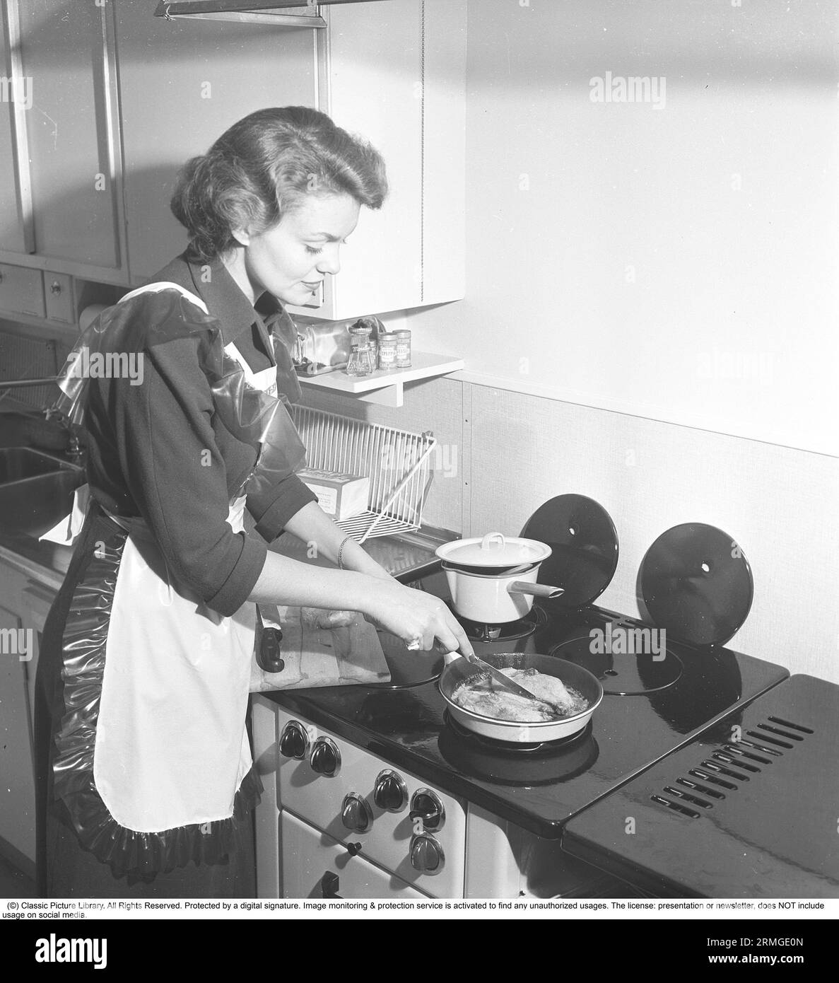 In der Küche der 1950er Das Innere einer Küche und eine junge Frau, die am Küchenherd steht und etwas kocht, das schmeckt. Sie trägt eine Plastikschürze. Sie ist Haide Göransson, 1928–2008, Modemodel und Schauspielerin. Schweden 1950 Kristoffersson ref AU23-3 Stockfoto