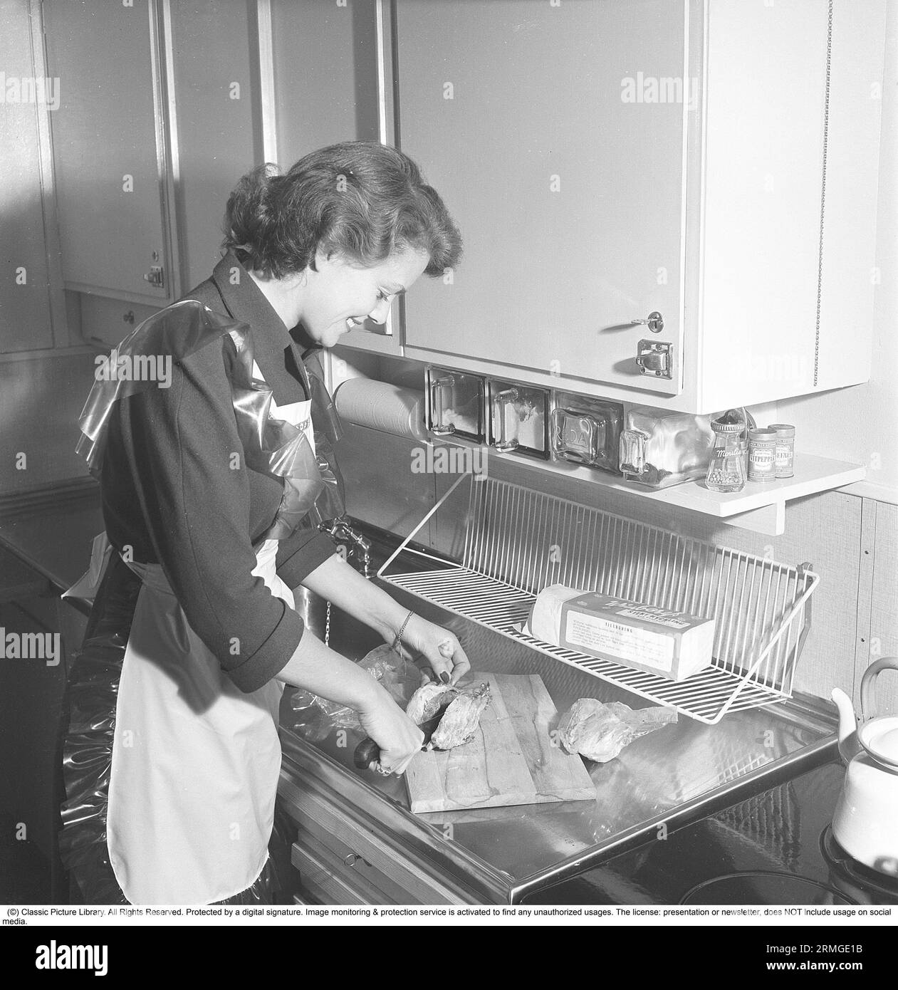 In der Küche der 1950er Das Innere einer Küche und eine junge Frau, die am Spülbecken steht und die Zutaten zubereitet. Sie trägt eine Plastikschürze. Sie ist Haide Göransson, 1928–2008, Modemodel und Schauspielerin. Schweden 1950 Kristoffersson ref AU23-2 Stockfoto