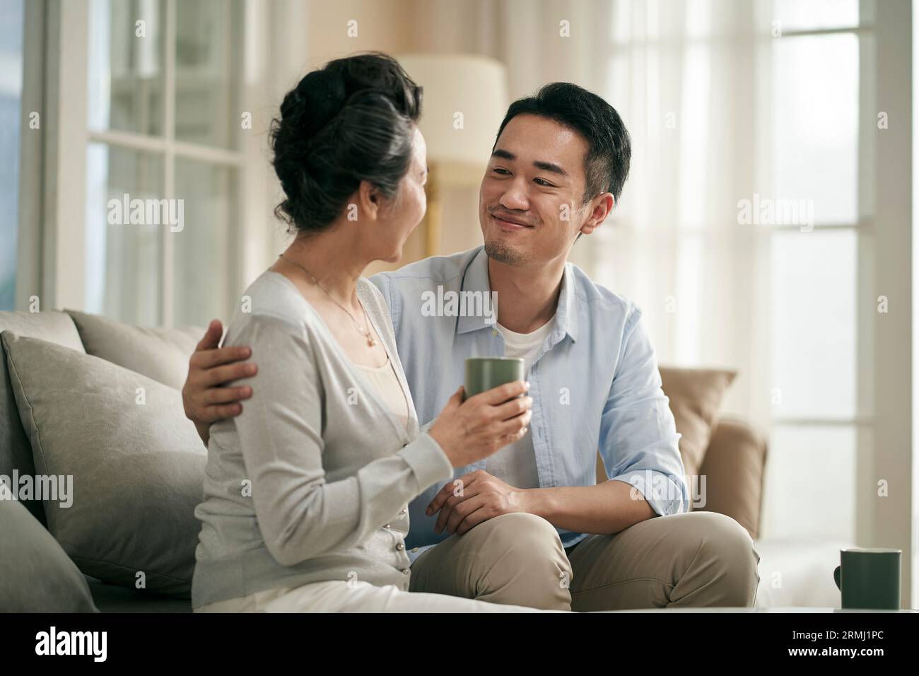 asiatischer erwachsener Sohn, der zu Hause auf dem Sofa sitzt und sich unterhält, mit älterer Mutter glücklich und lächelnd Stockfoto