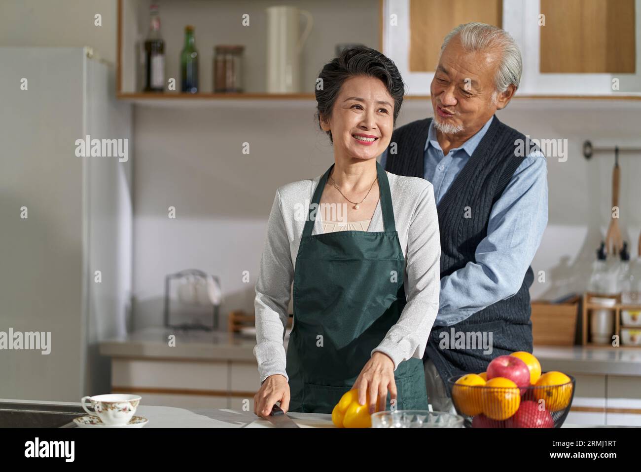 Liebender älterer asiatischer Mann Ehemann, der Frau hilft, die Schürze in der Küche zu Hause zu binden Stockfoto