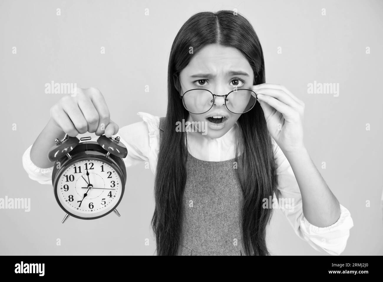 Teen Student Mädchen halten Uhr isoliert auf gelbem Hintergrund. Zeit zur Schule. Teenager-Kind mit Wecker, der die Zeit anzeigt, spätes Erwachen. Überrascht Stockfoto