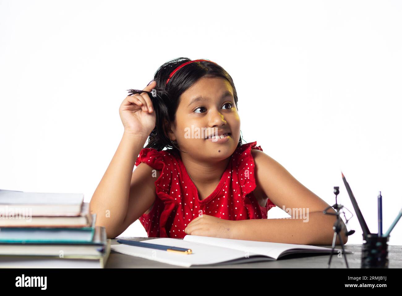 Ein hübsches indisches Mädchen, das denkt und nach oben schaut, während es am Lerntisch lernt, mit Büchern auf weißem Hintergrund Stockfoto
