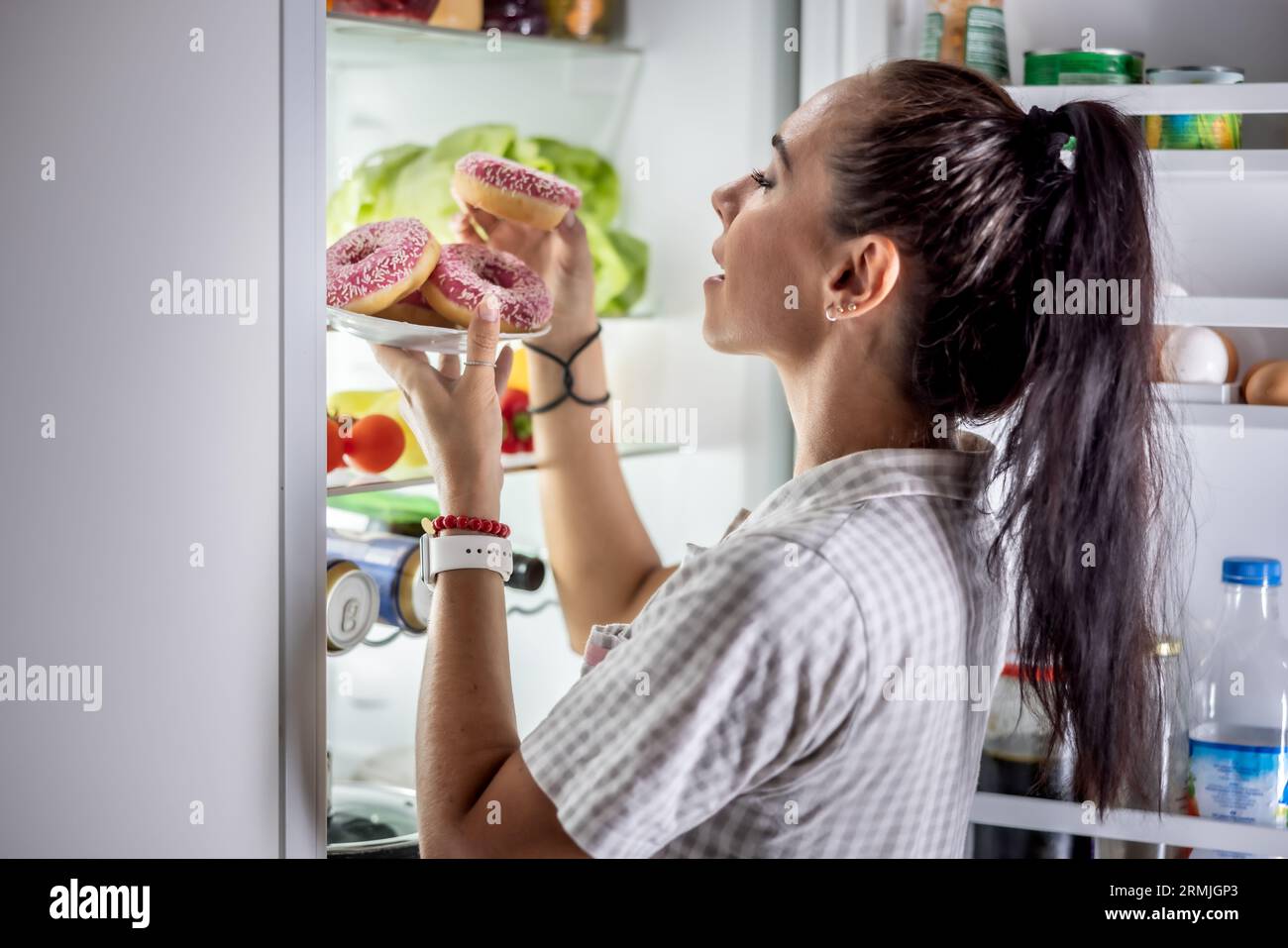 Eine junge hungrige Frau ist kurz davor, am späten Abend am offenen Kühlschrank süße Donuts zu essen. Stockfoto