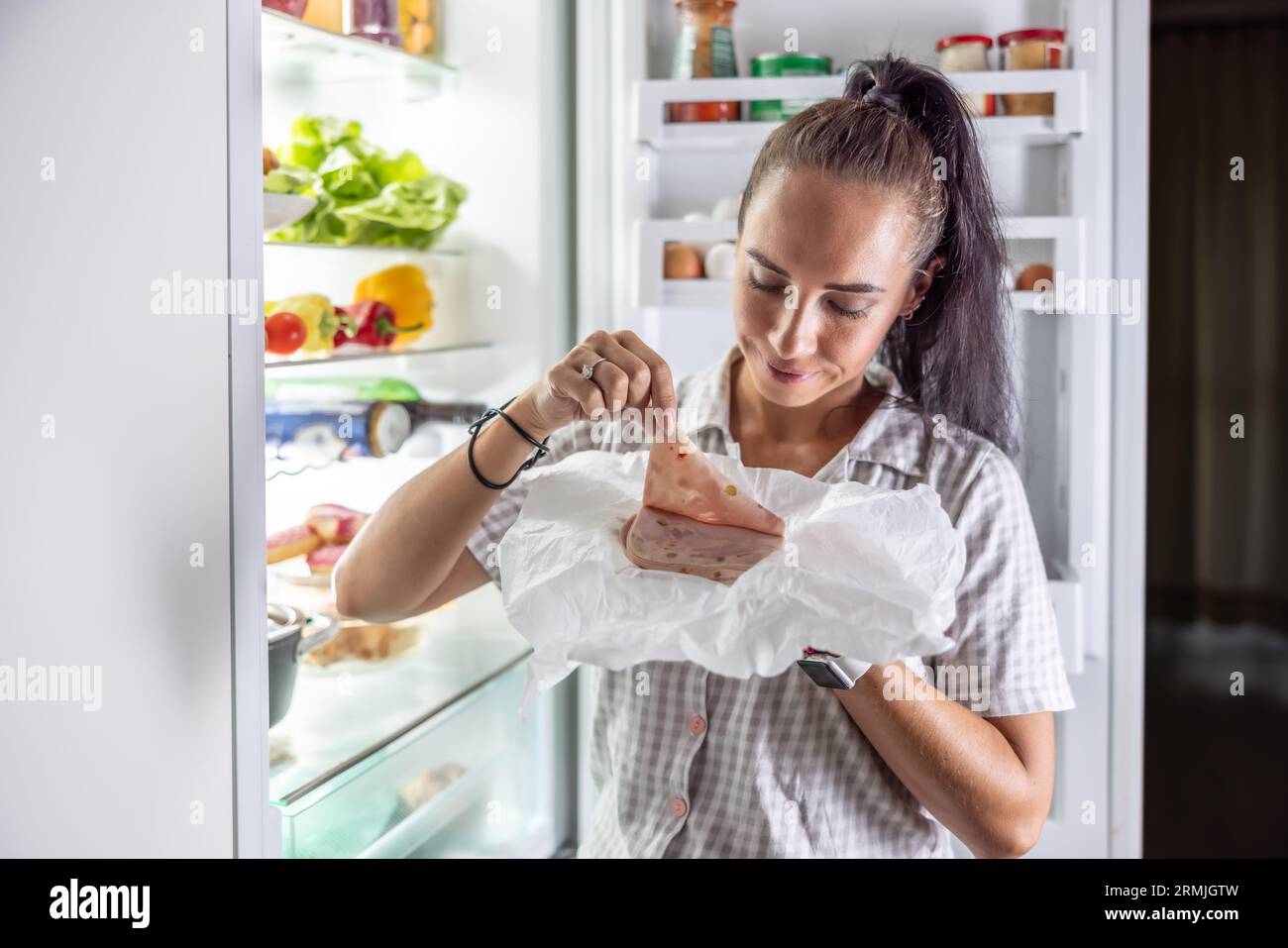 Sehr hungrige Frau im Pyjama, die abends am Kühlschrank Prosciutto genießt. Stockfoto