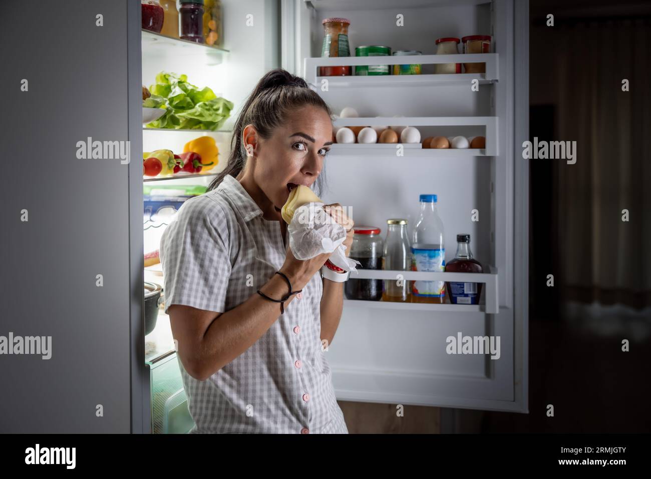Verängstigte hungrige Frau im Pyjama isst nachts Käse im Kühlschrank. Stockfoto