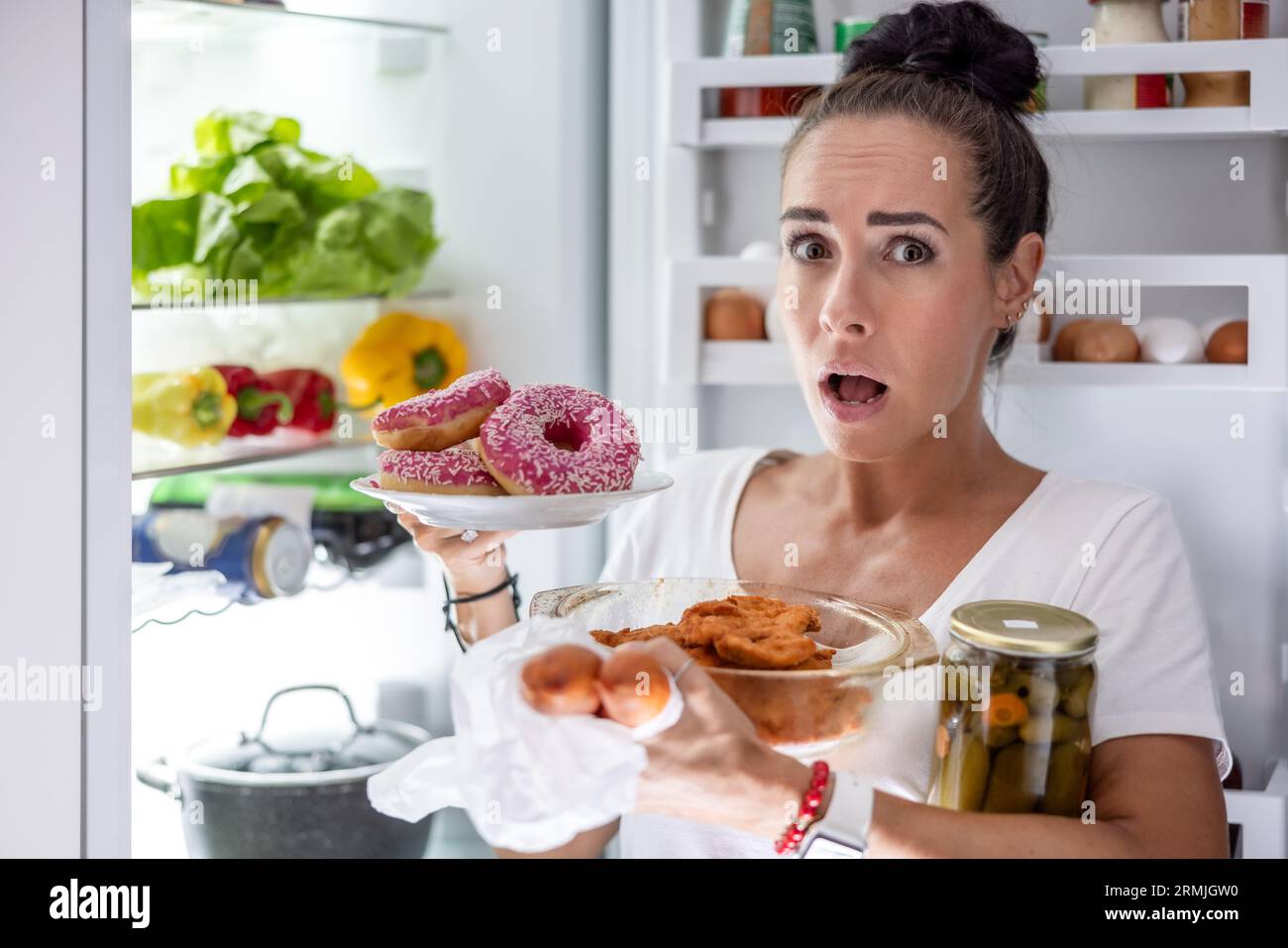 Am Freitagabend wählt eine sehr hungrige Frau im Pyjama Snacks, Steaks, Donuts und eine Dose Gurken aus dem Kühlschrank. Stockfoto