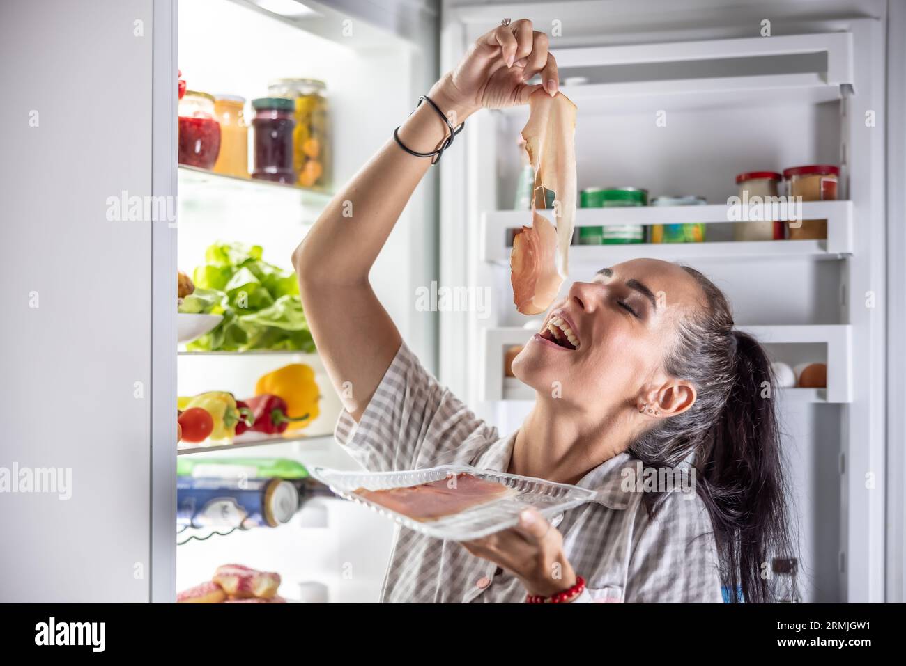 Sehr hungrige Frau im Pyjama, die abends am Kühlschrank Prosciutto genießt. Stockfoto
