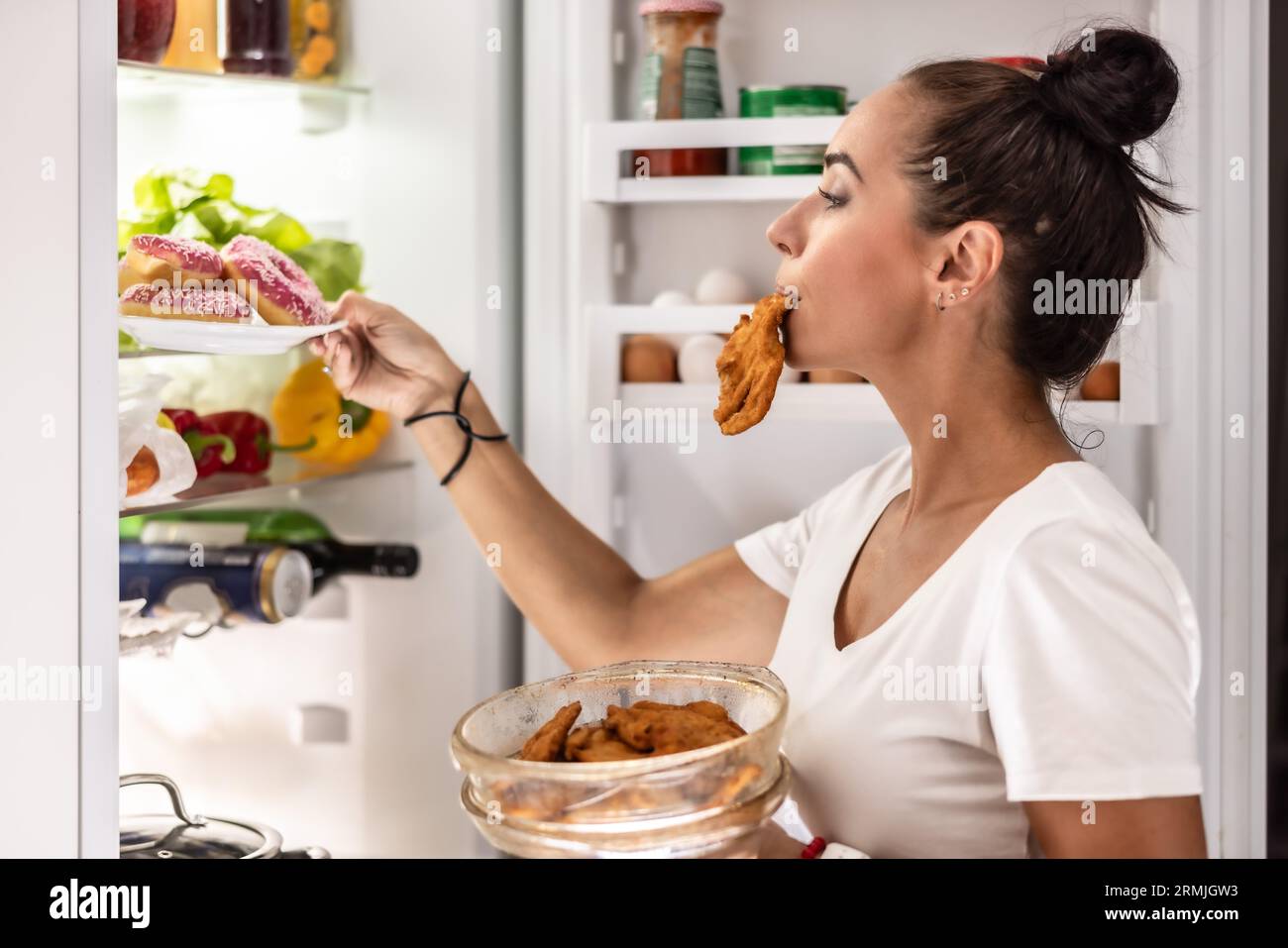 Eine sehr hungrige Frau im Schlafanzug wählt eine Schüssel Schnitzel und Donuts aus dem Kühlschrank. Stockfoto