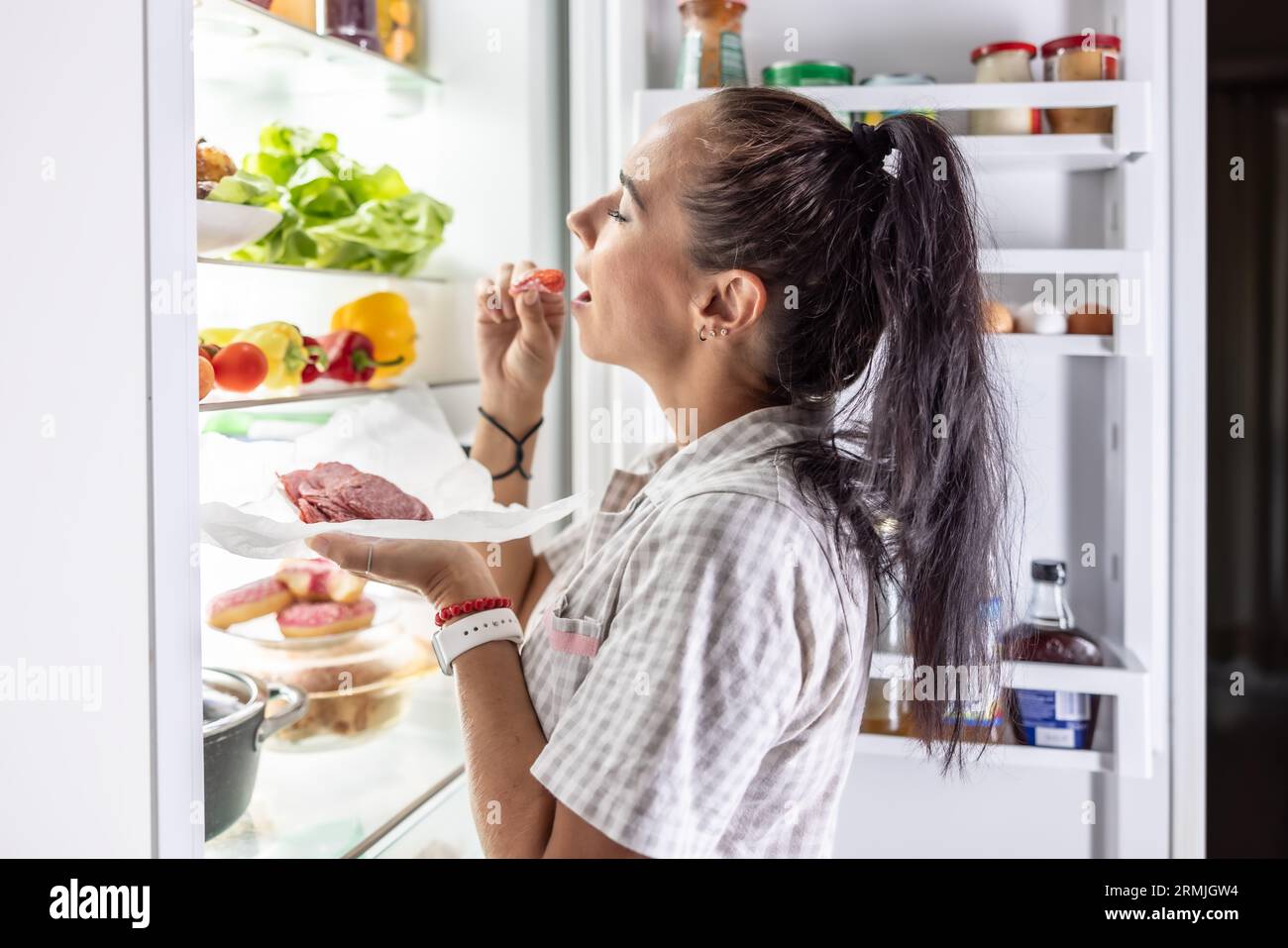 Sehr hungrige Frau im Pyjama, die nachts am Kühlschrank Salami genießt. Stockfoto